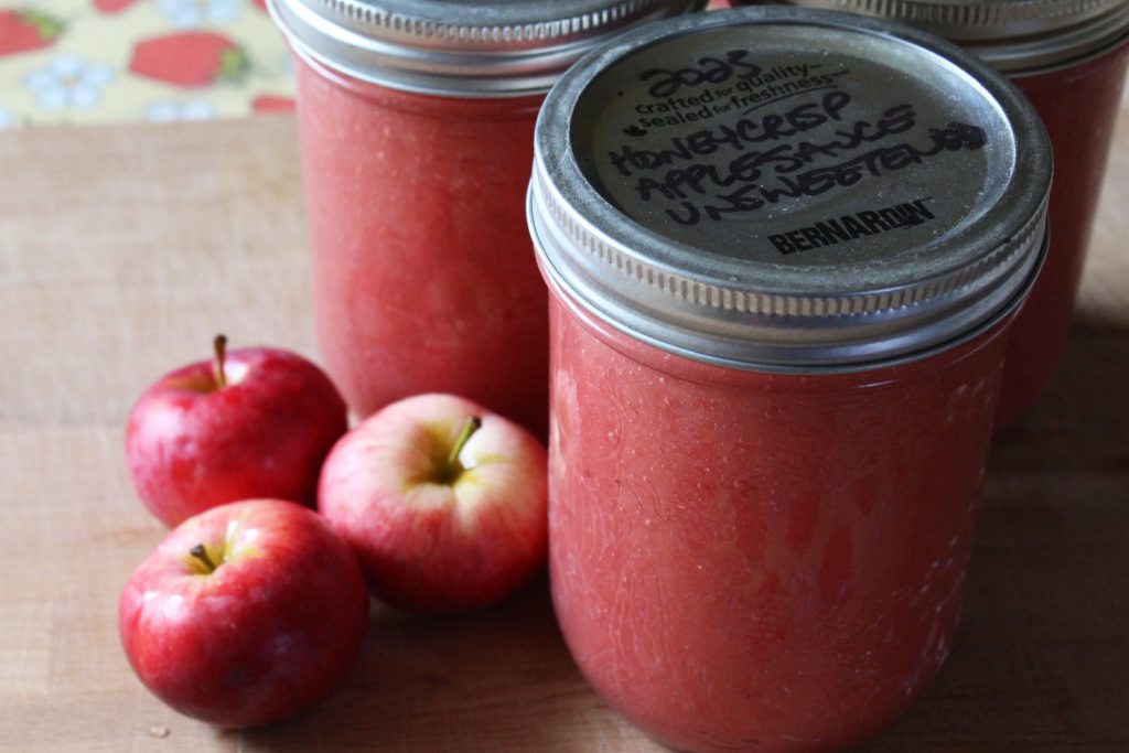 Jars of pink applesauce, ready to go in the pantry, with a few small red apples for decoration.